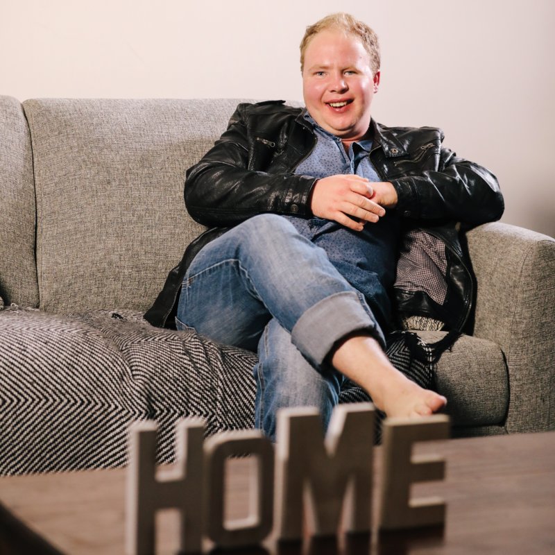 A man called Shane, sitting on his couch with a decorative piece which says home in the front.